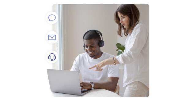 A smiling support agent wearing headphones sits at a laptop while a colleague points at the screen. On the left, floating icons for chat, email, and voice calls represent communication tools.